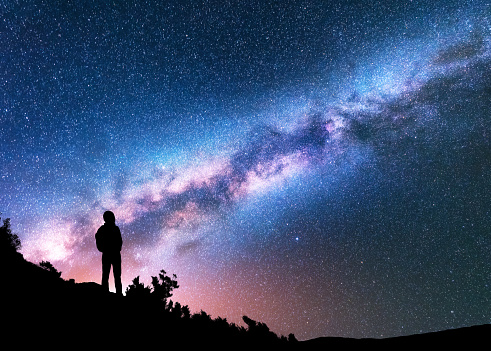 Silhouette of man with backpack on the hill against colorful Milky Way at night. Space background. Landscape with man, bright milky way, sky with stars. Beautiful galaxy. Travel. Starry sky. Universe