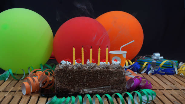 istock Chocolate birthday cake with five yellow candles extinguished on rustic wooden table with background of colorful balloons, gifts, plastic cups and streamers 915543820