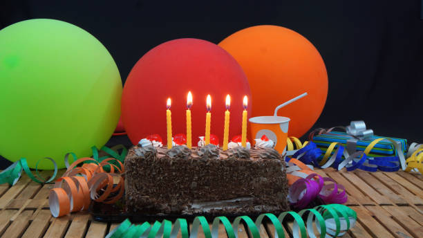 istock Chocolate birthday cake with five yellow candles burning on rustic wooden table with background of colorful balloons, gifts, plastic cups and streamers 915543826