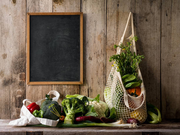 légumes frais du marché suspendus dans un sac en coton réutilisable à côté d’un tableau noir, sur un fond de mur vieille planche de bois, au-dessus d’une vieille table en bois avec d’autres légumes. - raw potato organic rustic bag photos et images de collection
