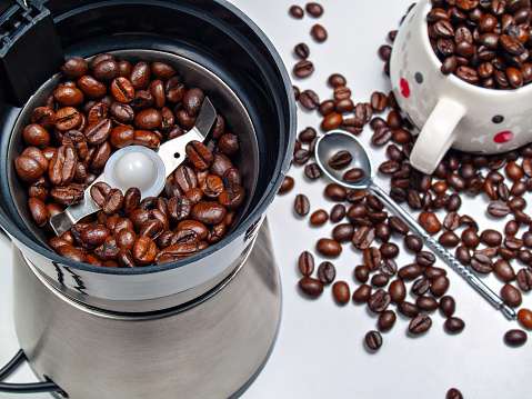 Grains of coffee in an electric coffee grinder, scattered grains on the table, a spoon and a cup of coffee beans