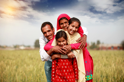 Cheerful farmer family standing in the field of wheat crop & smiling during springtime at the time of sunrise.