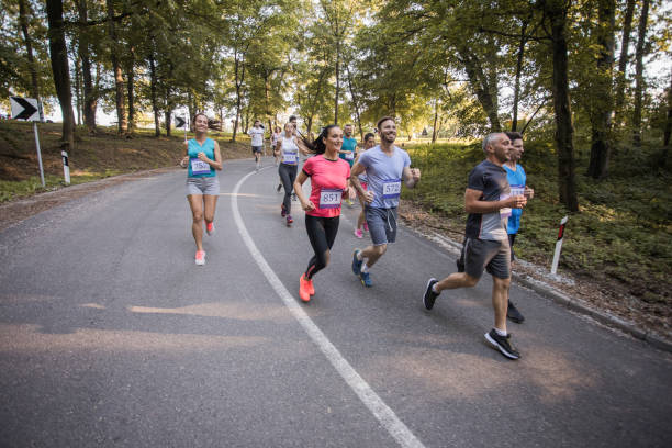 Large group of people running a marathon on asphalt road in nature. Large group of smiling athletes running a marathon through nature. distance running stock pictures, royalty-free photos & images
