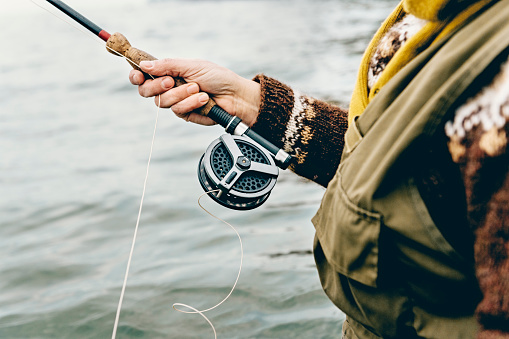 Close up portrait of a  fly fisherman's hands as she works the line and reel. She is wearing a green cotton gillet,waders, a patterned sweater and a mustard coloured scarf. Colour, horizontal format photographed against a calm sea with lots of copy space. Photographed on location at Nordfeld Strand on the island of Møn in Denmark.