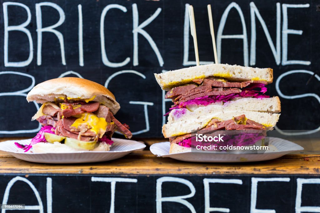 Close up of salt beef and mustard sandwiches with Brick Lane sign in background Close up color image depicting salt beef and mustard sandwiches in a row, for sale at a food market. In the background, blurred out of focus, is a sign saying Brick Lane - a street in London famous for its multitude of curry houses. Room for copy space. Brick Lane - Inner London Stock Photo