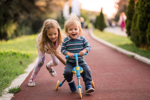 Happy boy having fun on tricycle while his sister is pushing him. Little girl helping her small brother with riding a tricycle in the park. tricycle stock pictures, royalty-free photos & images