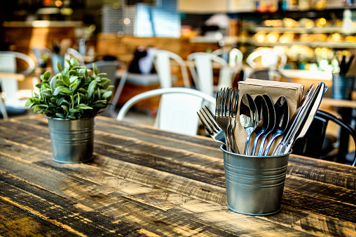 A warm cafe interior with wooden table, cutlery, pot plant and food counter in the background