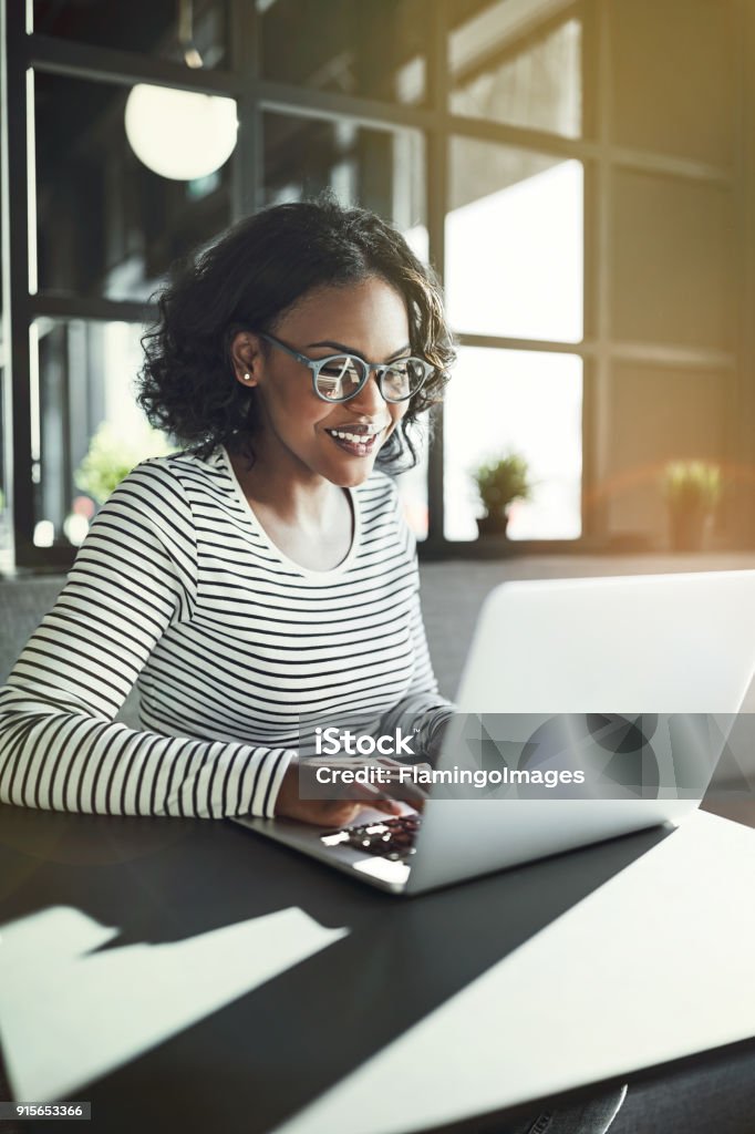 Sonriente joven mujer africana sentada en una mesa de trabajo en línea - Foto de stock de Ordenador portátil libre de derechos