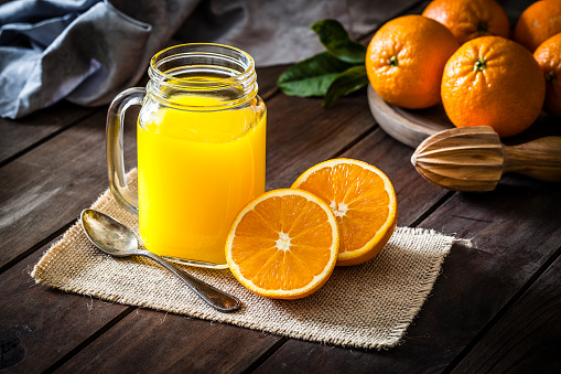 Orange juice glass jar shot on rustic wooden table. The jar is on a burlap cloth and two orange halves are beside it. An old metal spoon and a wooden juicer complete the composition. A round wooden tray with fresh oranges is at the top-right corner of an horizontal frame. Predominant colors are orange and brown. DSRL studio photo taken with Canon EOS 5D Mk II and Canon EF 100mm f/2.8L Macro IS USM