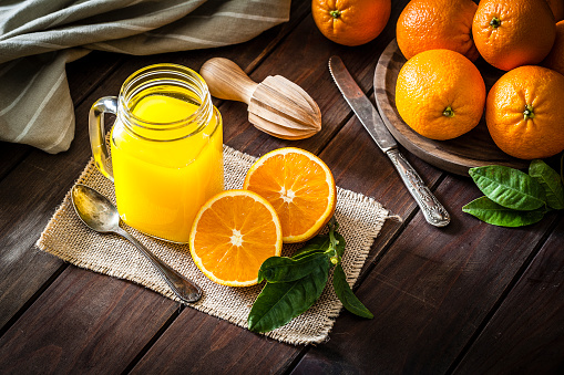 High angle view of an orange juice glass jar shot on rustic wooden table. The jar is on a burlap cloth and two orange halves are beside it. An old metal spoon and a wooden juicer complete the composition. A round wooden tray with fresh oranges is at the top-right corner of an horizontal frame. Predominant colors are orange and brown. DSRL studio photo taken with Canon EOS 5D Mk II and Canon EF 100mm f/2.8L Macro IS USM