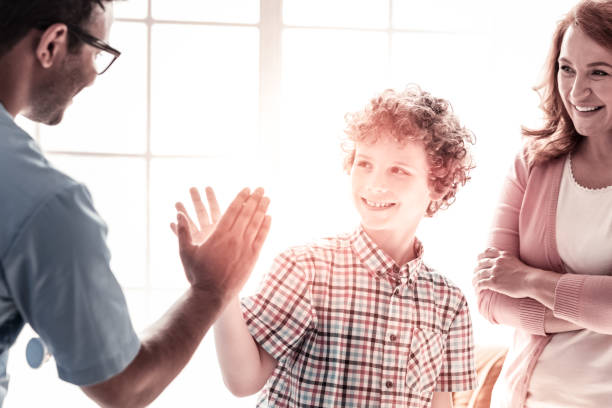 Positive minded boy and medical worker high fiving Be careful. Healthy smiling kid giving a high five to his friendly doctor after getting better and treating at a hospital. high minded stock pictures, royalty-free photos & images