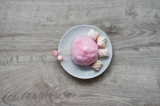 Composition of pink sweet cotton wool with hearts on wooden sticks and colorful soufflé on a wooden background. Sweet entertainment for children and their parents. Culinary delights for St. Valentine's Day.