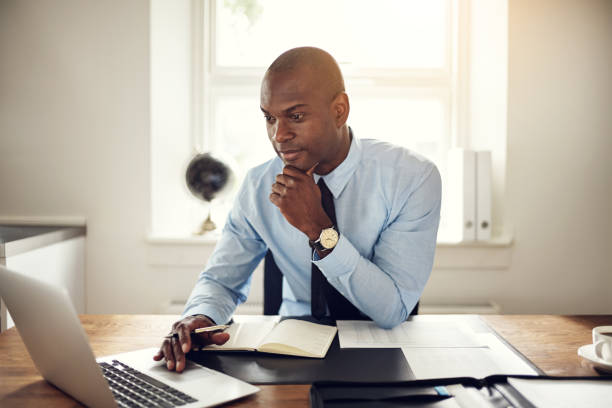 Young businessman working on a laptop in an office Young African business executive wearing a shirt and tie sitting at his desk in an office working online with a laptop black man laptop stock pictures, royalty-free photos & images