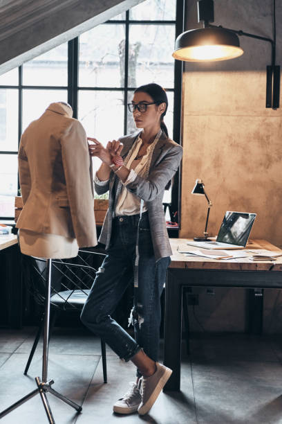 Successful fashion designer. Full length of serious young woman in eyewear using sewing needles for sewing a jacket on mannequin while standing in her workshop fashion designer photos stock pictures, royalty-free photos & images