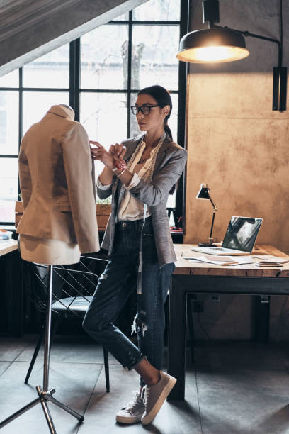 Successful fashion designer. Full length of serious young woman in eyewear using sewing needles for sewing a jacket on mannequin while standing in her workshop woman stitching stock pictures, royalty-free photos & images