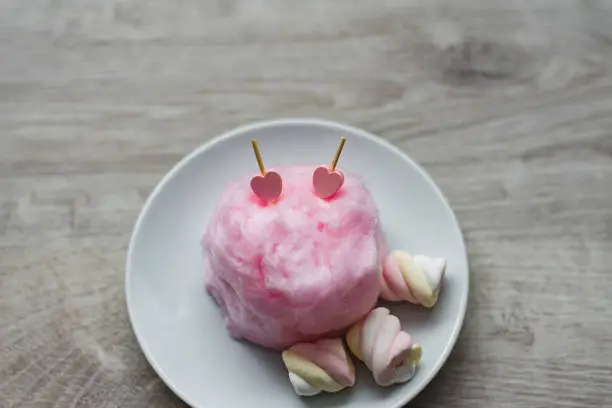 Composition of pink sweet cotton wool with hearts on wooden sticks and colorful souffle on a wooden background. Sweet entertainment for children and their parents. Culinary delights for St. Valentine's Day.