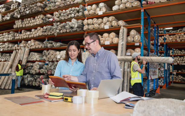 Man and woman looking at brochure in carpet manufacturing warehouse Mature and and young Asian colleague talking and discussing designs, factory workers in background carpet factory photos stock pictures, royalty-free photos & images