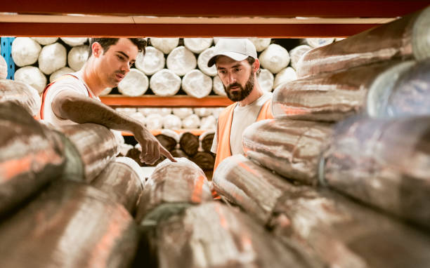 Two men discussing carpets in warehouse storage facility Male colleagues working on factory floor looking at rolled up carpets wrapped in plastic carpet factory stock pictures, royalty-free photos & images