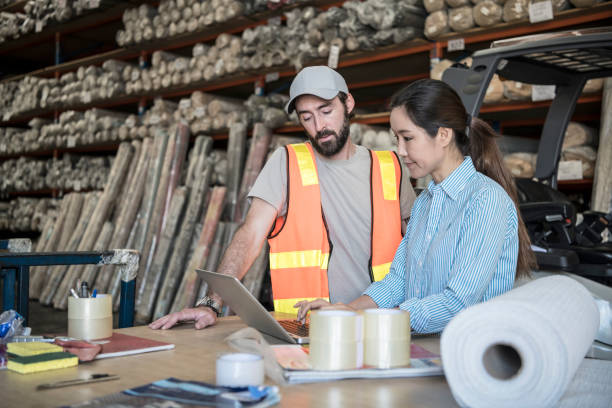 Woman using laptop in warehouse with male employee watching Two people working in carpet factory, female manager on computer, mid adult man wearing hi vis carpet factory stock pictures, royalty-free photos & images