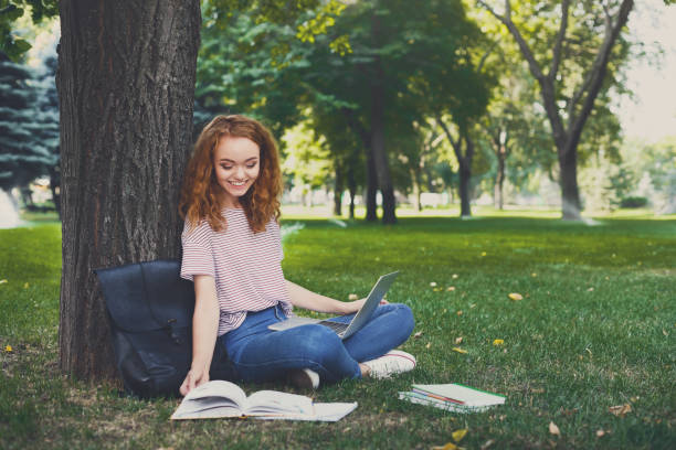 istock Redhead girl outdoors reading book and using laptop sitting under a tree 915704274