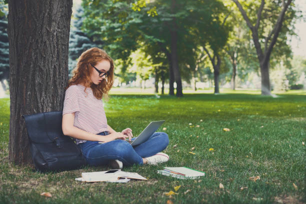istock Young redhead girl in eyeglasses using laptop sitting under a tree 915704310