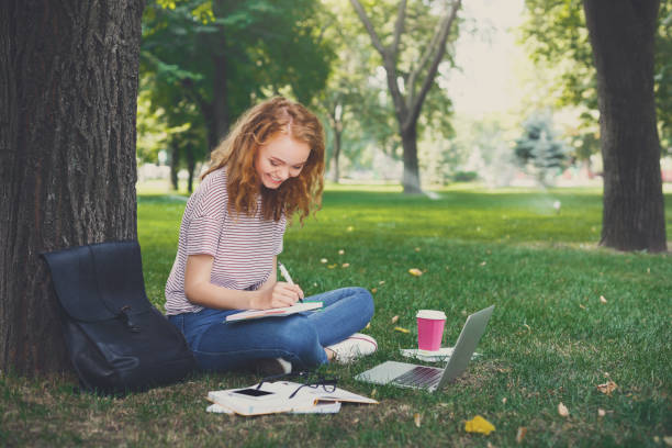 istock Young redhead girl writing in notebook sitting under a tree 915704334
