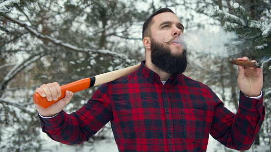 Bearded man with axe in winter outdoors, smoking cigar. Shallow depth of field