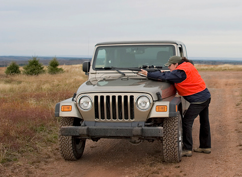 A woman hunter aims a rifle over the hood of a 4x4.