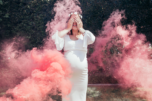 Happy pregnant woman covering her face with smoke grenade outdoors. Happy young expecting mother standing outside with smoke grenade.