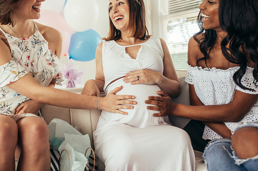 Group of smiling young female friends touching tummy of pregnant woman. Multiracial female friends having fun at baby shower.