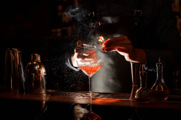 Bartender in black apron and blue shirt setting fire to sweet cocktail in bocal at a bar counter