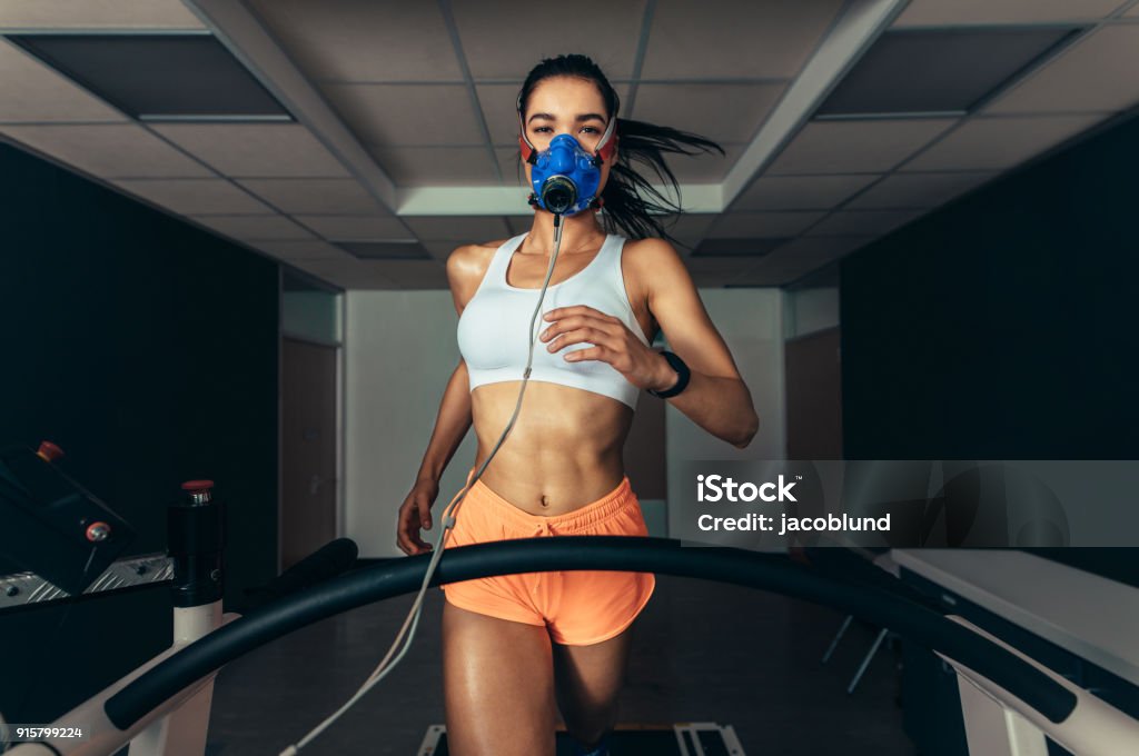 Sportswoman with mask running on treadmill Sportswoman with mask running on treadmill. Female athlete in sports science lab measuring her performance and oxygen consumption. Treadmill Stock Photo