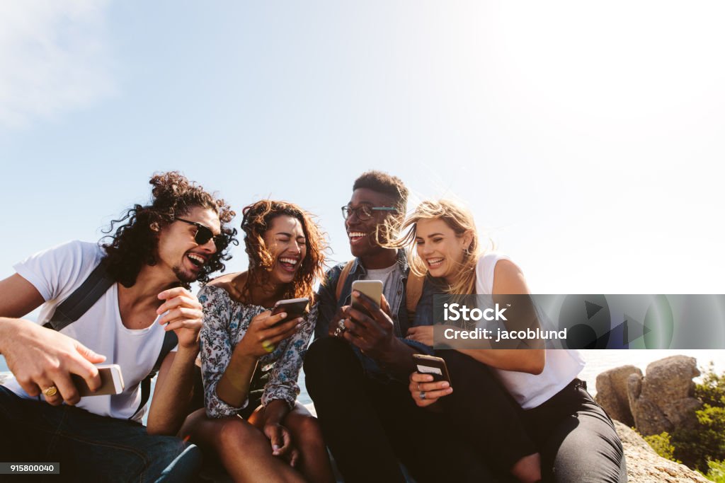 Group of people having fun on their holidays Young group of people sitting on top of mountain using smart phones and smiling. Diverse friends enjoying a day out. Friendship Stock Photo