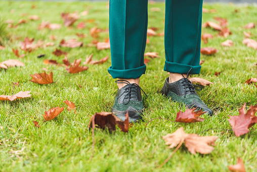 cropped view of female legs in green pants and brogue shoes standing on lawn with autumn foliage