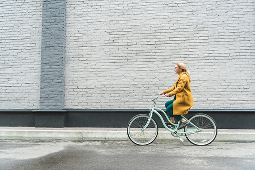 fashionable woman in yellow coat riding bicycle with grey wall on background
