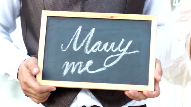 Wedding concept. Will you marry me question handwritten on blackboard shown by young men.