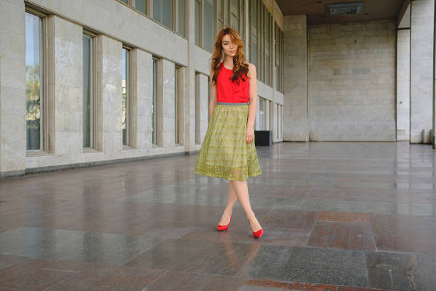 Stylish curly-haired girl in lace skirt, red shoes and blouse, posing with seductive face expression in a big space with many windows.