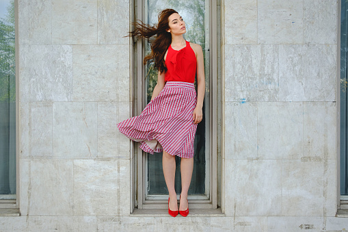 Gorgeous woman with flying hair and fluttering skirt in the wind posing while standing on a window like in a vitrine. Fashion, presenting clothes concept