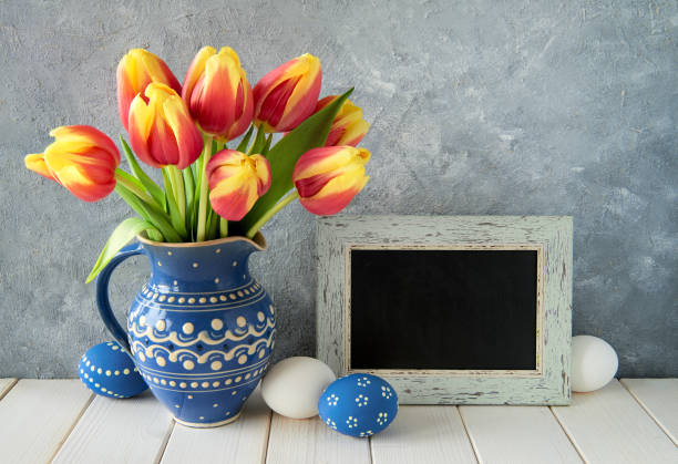istock Red-yellow tulips in blue ceramic pitcher with Easter eggs and a blackboard, text space 915874582