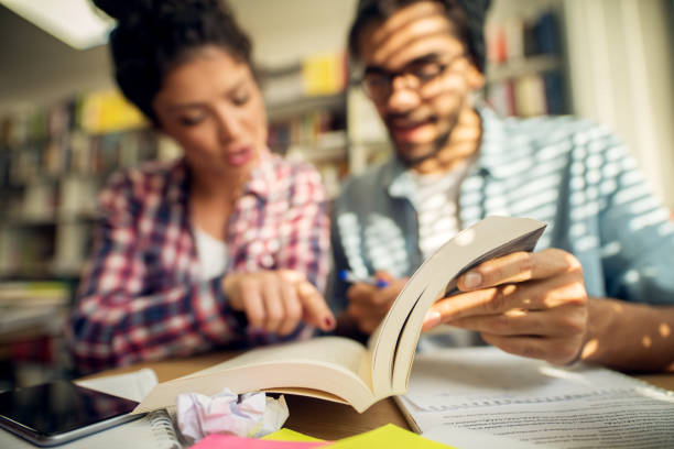 istock Concept of education, library, students, love and teamwork. Innovative productive young studying couple sitting in the library and learning together from a camera focused book. 915875612