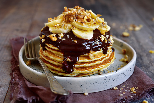 Stack of homemade delicious banana pancakes topped with chocolate sauce, banana slices and walnut on a plate on a dark wooden background.