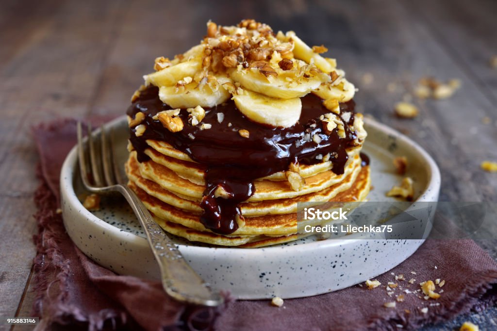Stack of homemade delicious banana pancakes topped with chocolate sauce, banana slices and walnut Stack of homemade delicious banana pancakes topped with chocolate sauce, banana slices and walnut on a plate on a dark wooden background. Pancake Stock Photo
