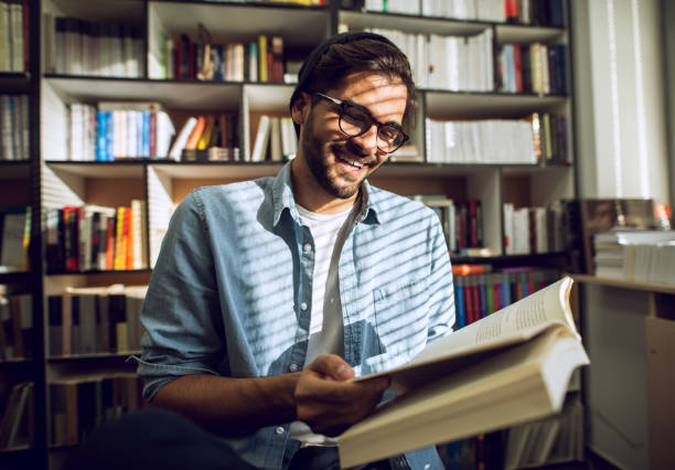 istock Handsome man hipster reading book at library smiling. Education at public. 915886230