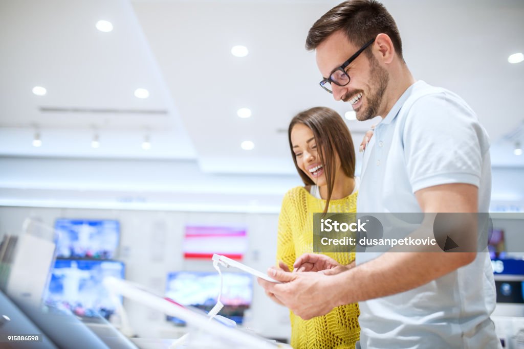 Affascinante giovane coppia d'amore sorridente che guarda su un tablet mentre acquista in un negozio di tecnologia. - Foto stock royalty-free di Negozio