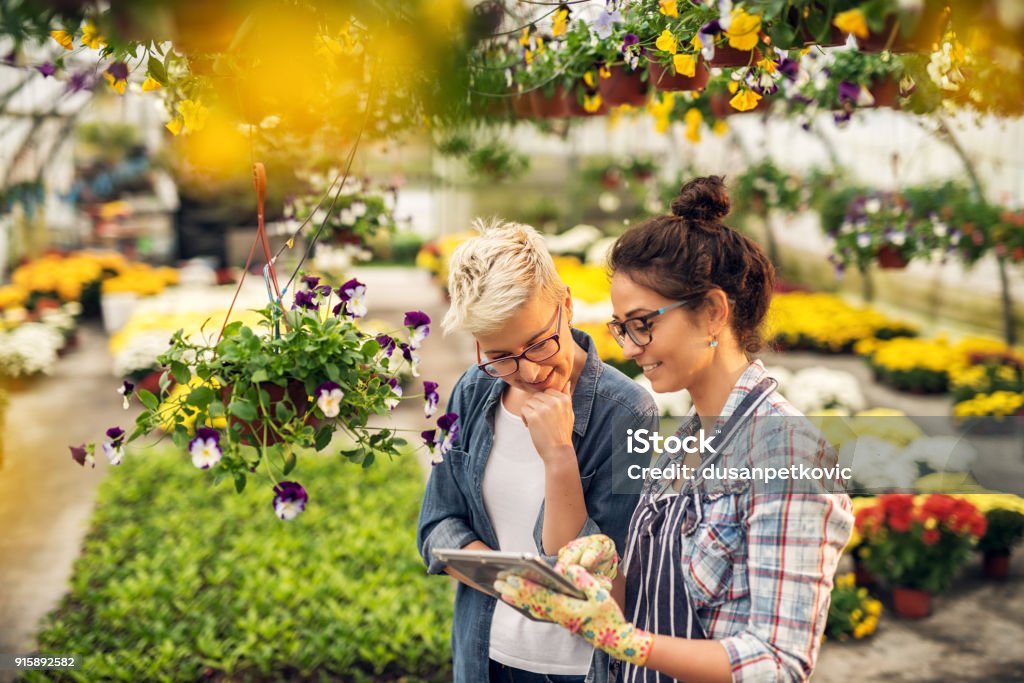 Adorable charming professional modern florist woman showing a list of flowers on a tablet to the curious attractive blonde female customer in a greenhouse. Gardening Stock Photo