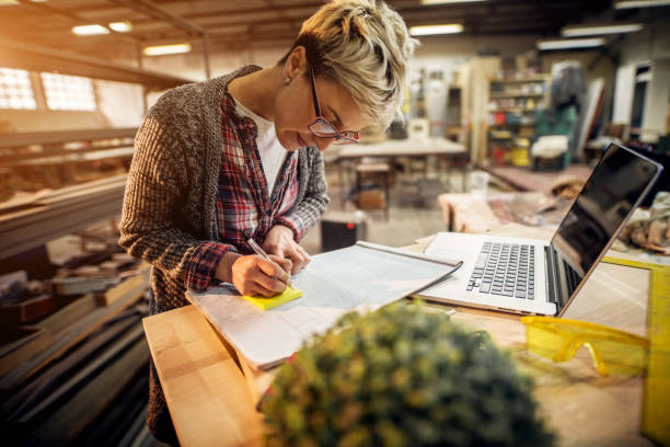 istock Hardworking professional engineer short hair woman writing on notes while working on a laptop and blueprint in the sunny workshop. 915904484