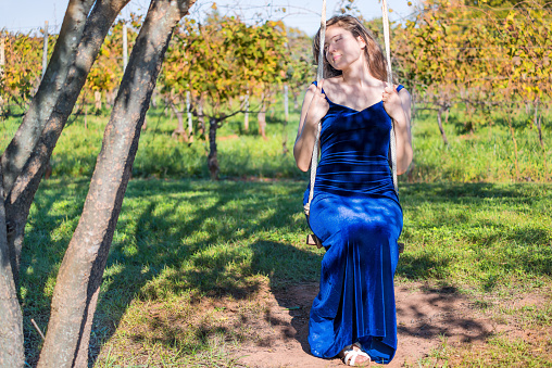 Elegant young woman in blue velvet dress by vineyard winery grapevine leaves green in Virginia grapes, front smiling happy sitting on tree swing