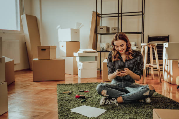 Young woman moving to a new apartment. Typing a message while sitting down on a carpet