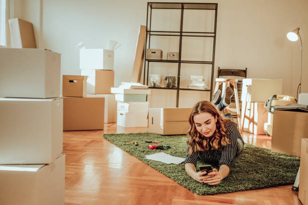 istock Young woman moving to a new apartment. Typing a message while sitting down on a carpet 915917702