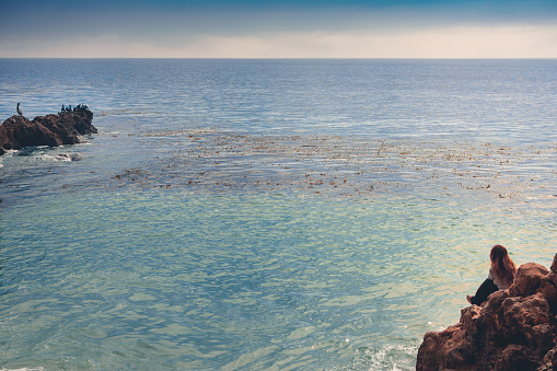 A woman is resting at the water's edge, as she sits alone on a ledge along the rocky coastline of Rancho Palos Verdes in Southern California. She's watching the birds in the distance. The waters are calm without much movement or waves. She is in her 40's and Asian.